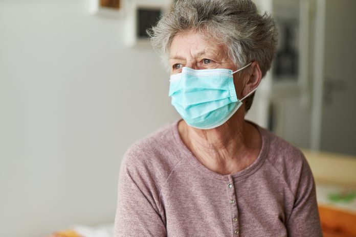 A senior citizen sits alone on her bed with a respirator or surgical mask